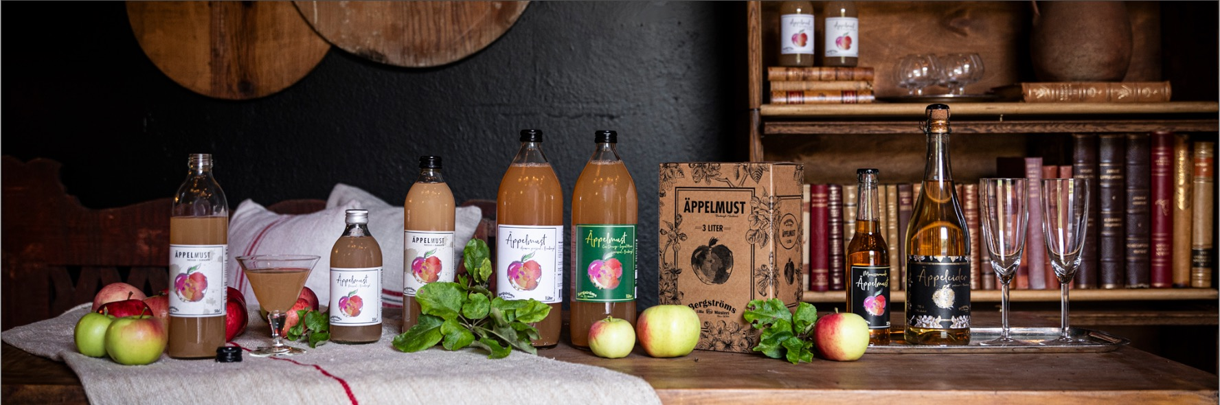 Sortiment av Bergströms drycker uppdukade på ett träbord — flaskor med äppelmust, bag-in-box, mousserande äppelmust och äppelcider mot en mörk vägg med bokhylla